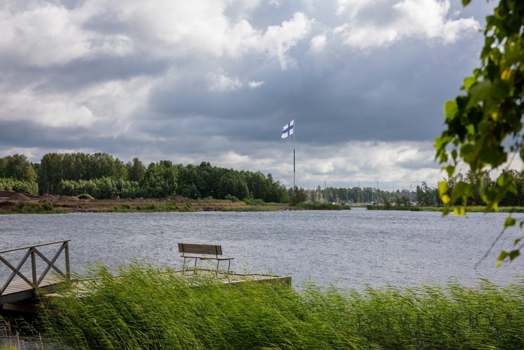 Edustalla kaislikko ja laituri sekä meri, taustalla Suomen lippu.